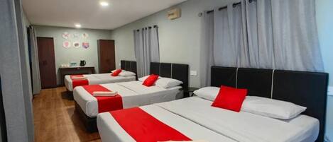 A hotel room with three beds, each with white bedding and red accents, wooden flooring, and grey curtains.