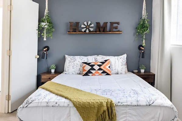 Un lit avec une literie à motifs, un panneau décoratif portant l’inscription « HOME » au mur et deux plantes suspendues.