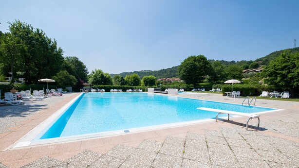 Ein rechteckiger Swimmingpool mit glasklar blauem Wasser, umgeben von weißen Liegestühlen und Sonnenschirmen, vor einer Kulisse aus üppigen grünen Bäumen und Hügeln.