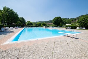 A rectangular swimming pool with a clear blue water surface, surrounded by white lounge chairs and umbrellas, set against a backdrop of lush green trees and hills. - Appartamento ai Colli (Padenghe sul Garda)