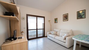 Ein Wohnzimmer mit weißem Sofa, einem kleinen Tisch, einem Fernseher auf einem Holzgestell und einer zur Außenseite führenden Tür.