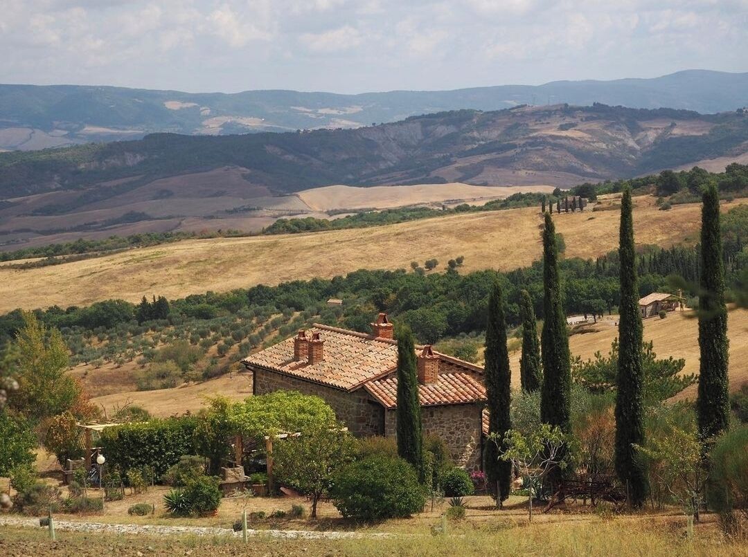 A rustic stone house with a tiled roof, surrounded by vineyards and cypress trees, set against rolling hills.