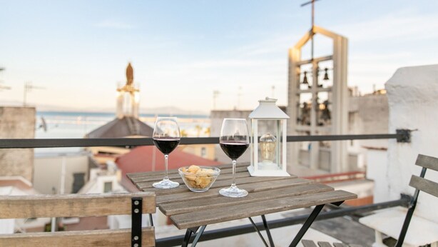 Ein Holztisch mit zwei Weingläsern und einer Schale mit Snacks auf einem Balkon mit Blick auf eine Stadtlandschaft, eine Kirche und den Meerblick.
