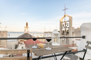 Une table en bois, avec deux verres à vin et un bol de collations, est placée sur un balcon qui offre une vue sur la ville, avec une église et la mer en toile de fond.