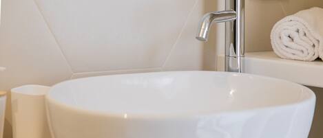 A white bathroom sink with a chrome faucet, a toilet paper holder, and a rolled towel on a shelf.
