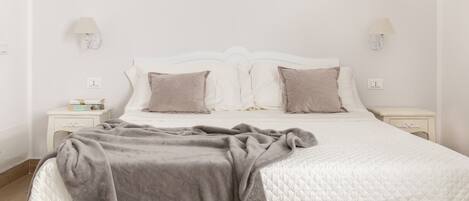 A neatly made bed with a quilted white duvet, two pillows, and a grey blanket. There are two bedside tables with lamps and a small bedside table with a book and a box.