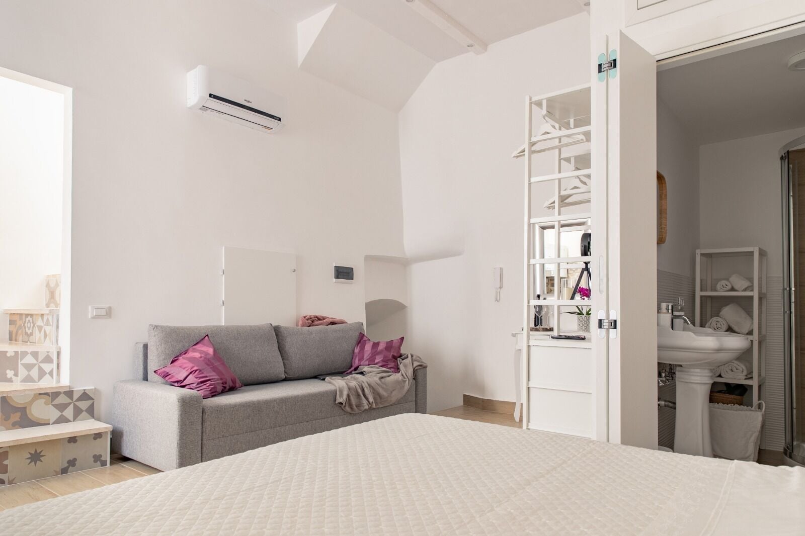 A bedroom with a bed, a grey sofa with pink cushions, a bedside table, and a white wardrobe.