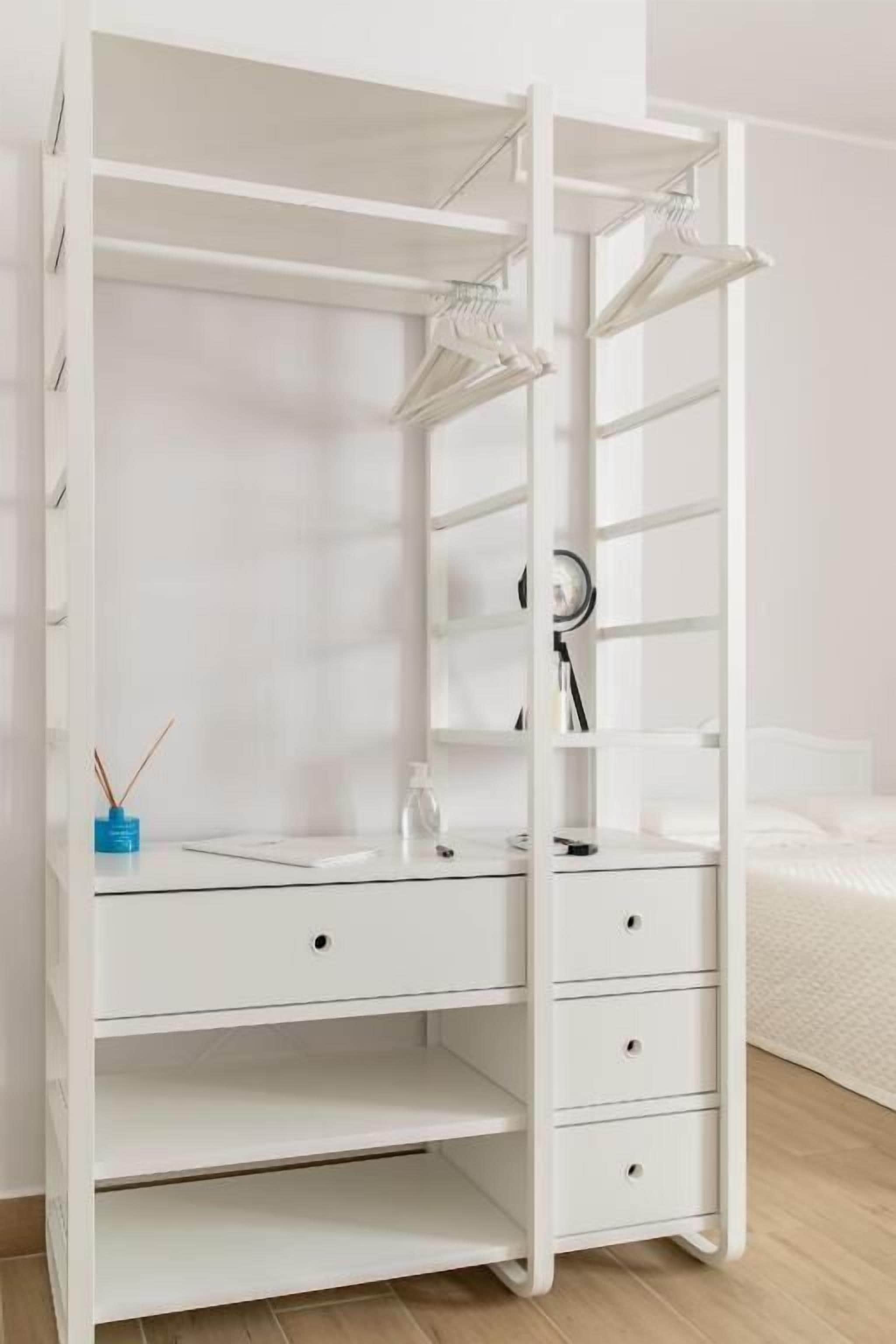 A white wardrobe with multiple shelves and drawers, a mirror, and a small desk area with a lamp and a blue container.