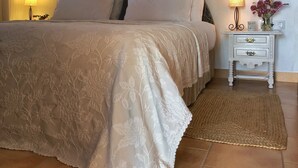 A neatly made bed with a floral-patterned bedspread, two bedside lamps, a small bedside table with a vase of flowers, and a woven rug on a tiled floor.