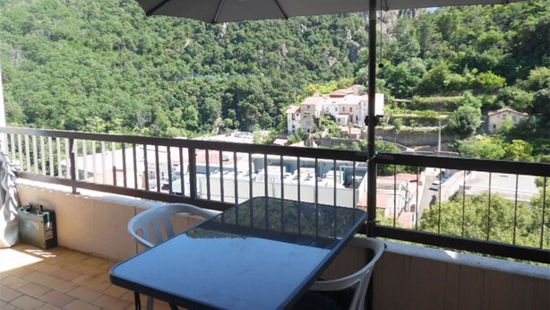 A balcony with a table and chairs, an umbrella, and a view of a green hillside with buildings. - Sunny Studio for 2 with Balcony, Private Parking, Wi-Fi, A/C, and Close to Thermes and City Center (Amélie-les-Bains-Palalda)