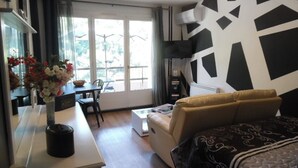 A modern living room with a sofa, a dining table with chairs, a small kitchen area, and a large window with a view of trees. - Sunny Studio for 2 with Balcony, Private Parking, Wi-Fi, A/C, and Close to Thermes and City Center (Amélie-les-Bains-Palalda)