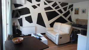 A living room with a modern geometric wall design, a white sofa, a dining table with a basket of fruit, and a bed with patterned bedding. - Sunny Studio for 2 with Balcony, Private Parking, Wi-Fi, A/C, and Close to Thermes and City Center (Amélie-les-Bains-Palalda)