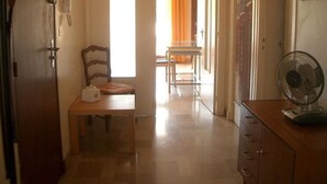 A hallway with a wooden desk, a chair, and a fan. There is a door on the left and a window with curtains on the right.