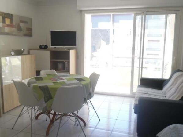 A modern living room with a round table, white chairs, a sofa, a TV on a stand, and a sliding glass door to the outside.