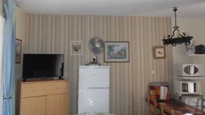 A living room with a television, a refrigerator, a microwave, a fan, and a dining table with chairs.