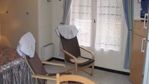 A room with two wooden chairs, a bed with a patterned cover, a radiator, a cabinet, and a window with curtains.