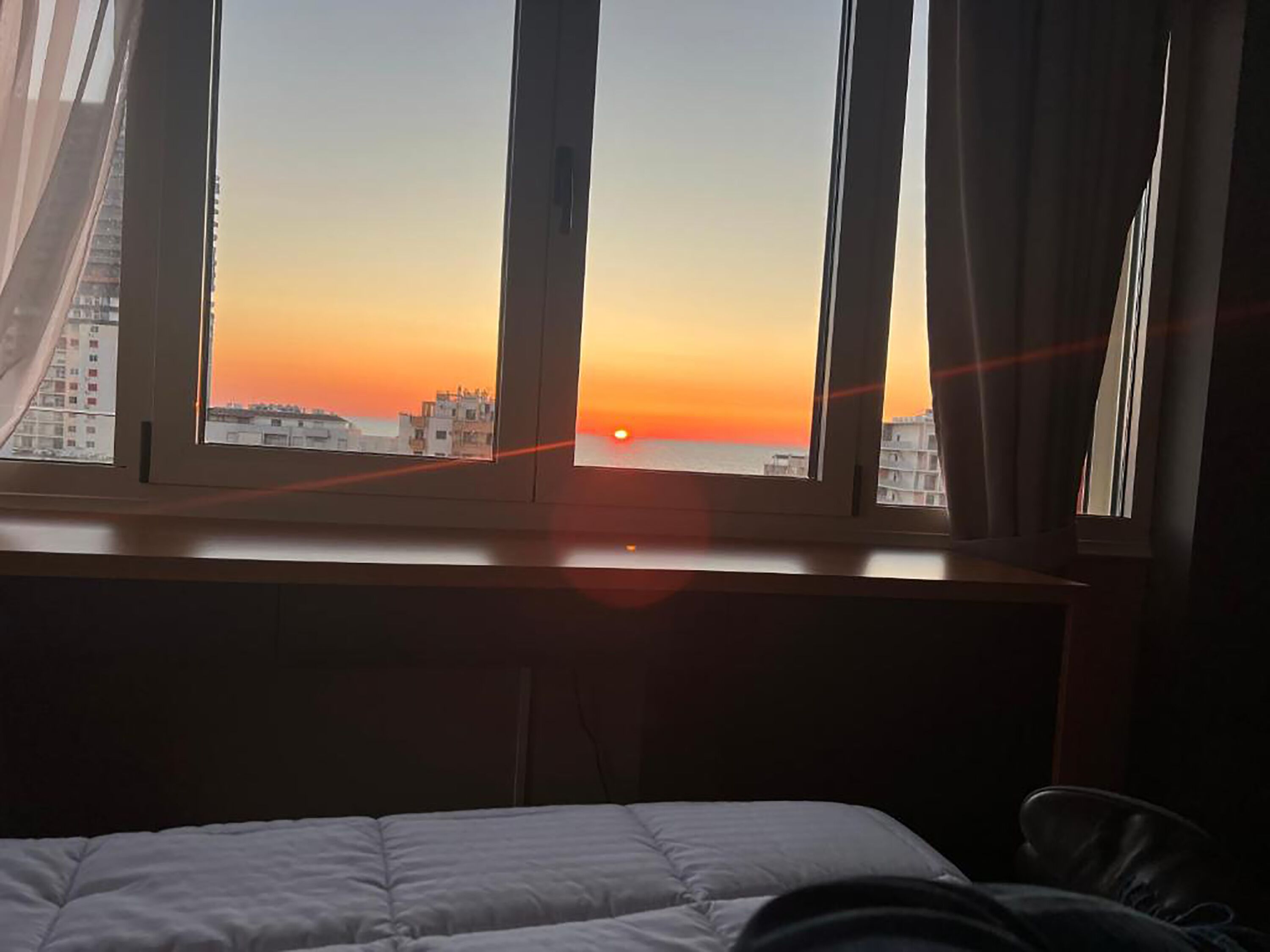 A hotel room with a bed, a window with curtains, and a view of the sunset over a cityscape.