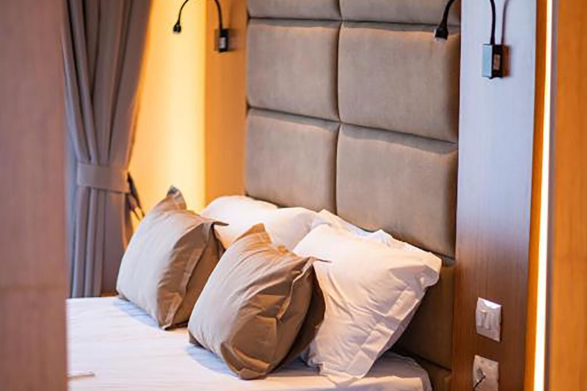 Een net opgemaakt bed met kussens, een nachtkastje met een telefoon, en een wandlamp.