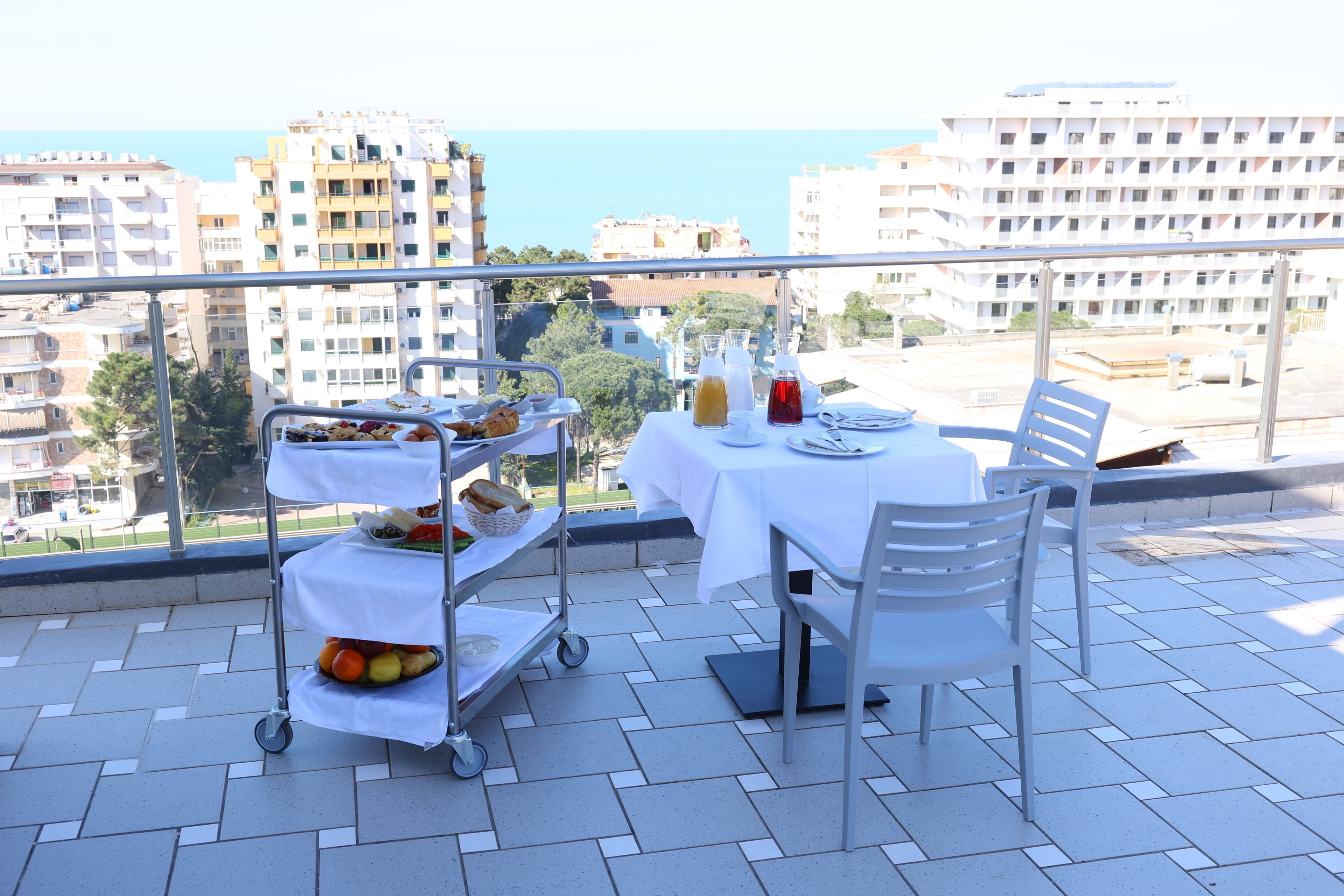 A balcony with a table set for two, a fruit cart, and a city view.