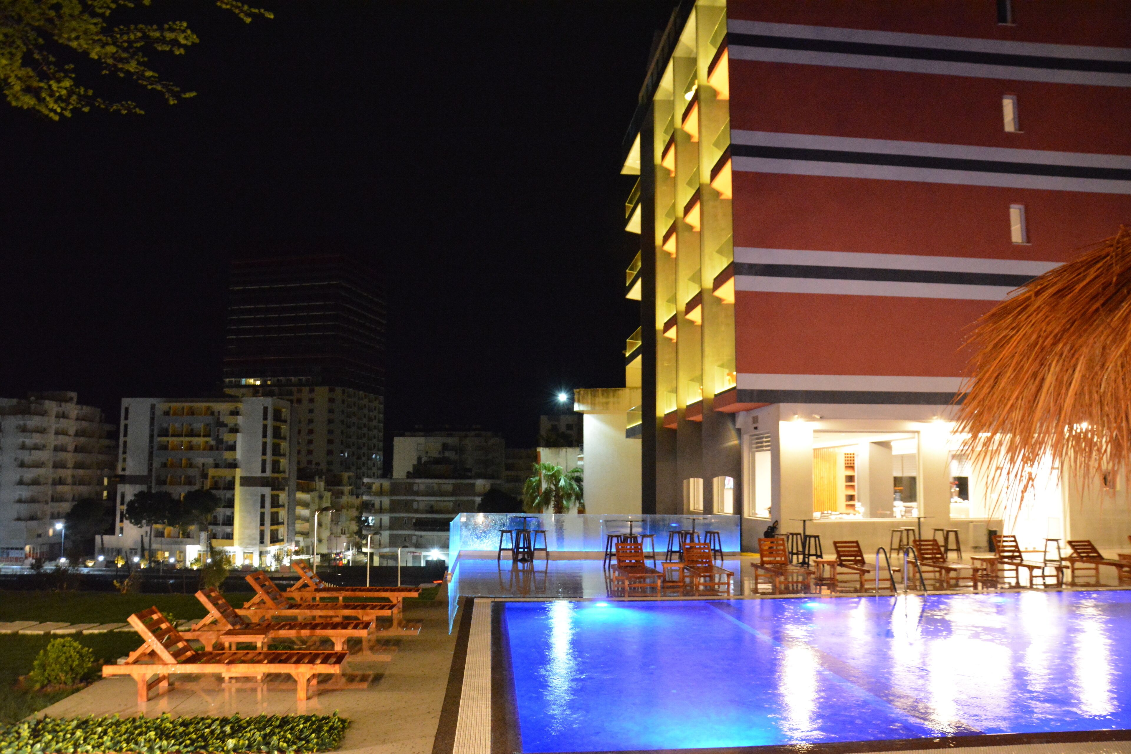 A nighttime view of a hotel pool area with lounge chairs and a building with a red and yellow facade.