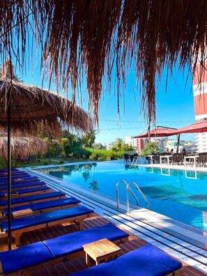 Un espace aménagé au bord de la piscine, avec des chaises longues, un parasol en chaume et une piscine à l’eau d’un bleu limpide.