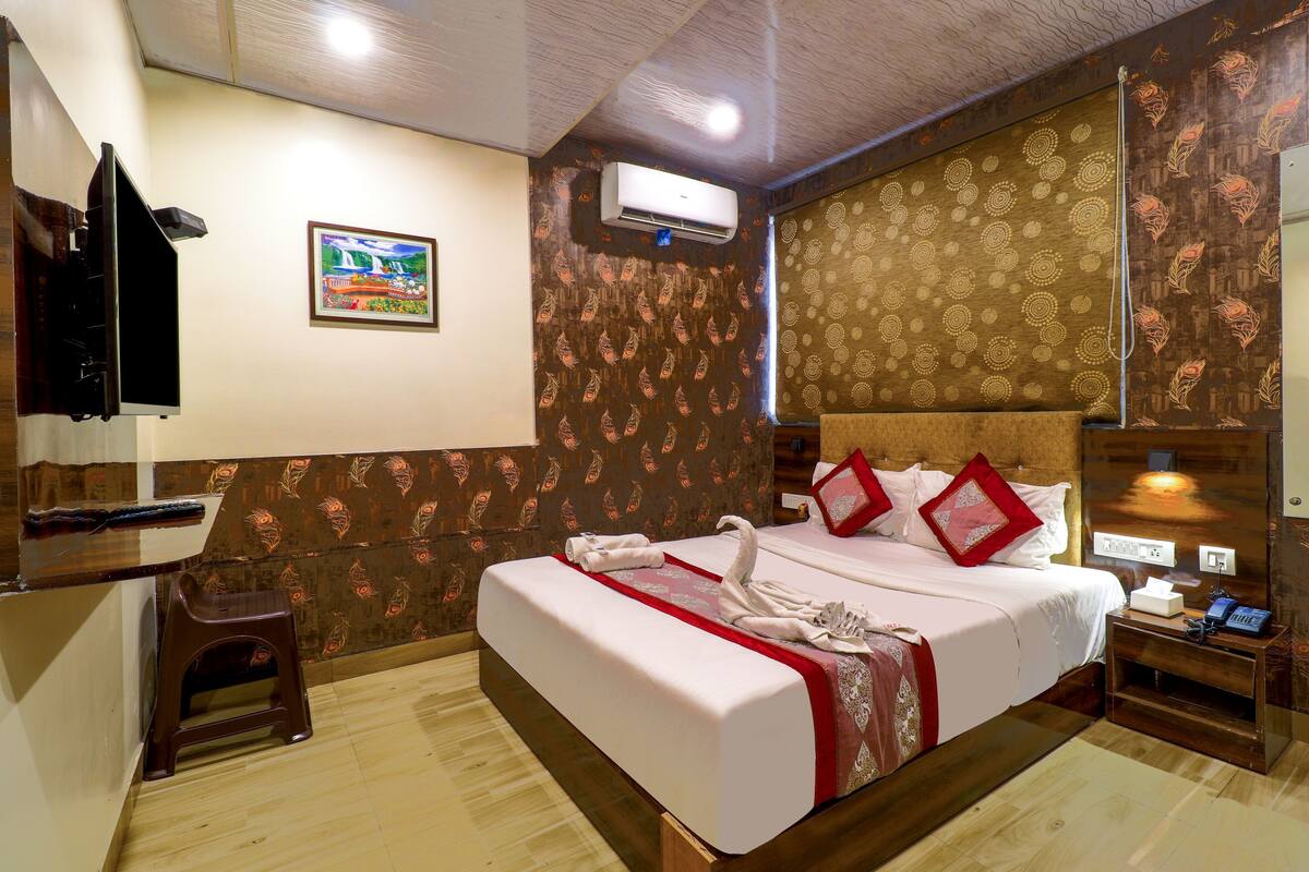 A hotel room with a bed, a small stool, a nightstand, an air conditioning unit, and a wall-mounted TV.