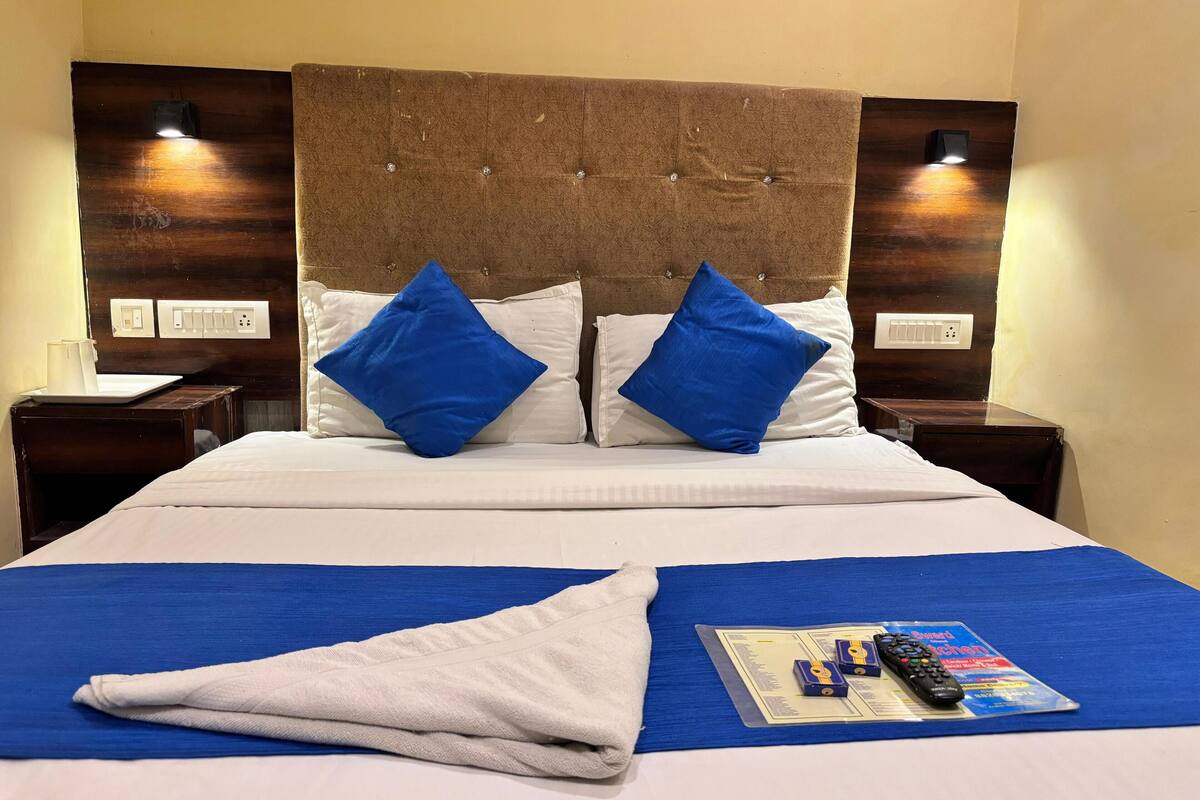 A hotel room with a large bed, two blue pillows, a white blanket, a remote control, and a magazine on a blue mat.