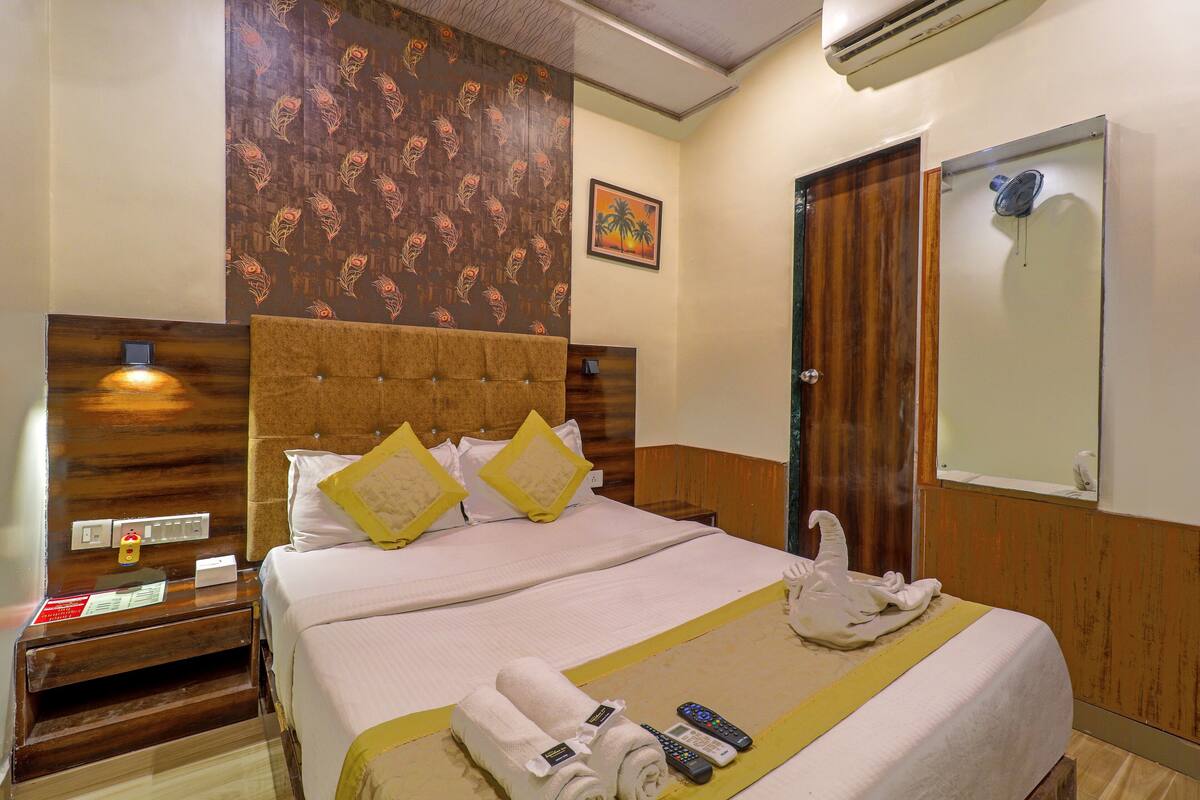 A hotel room with a large bed, a wooden headboard, a wall-mounted air conditioner, a mirror, and a framed picture.