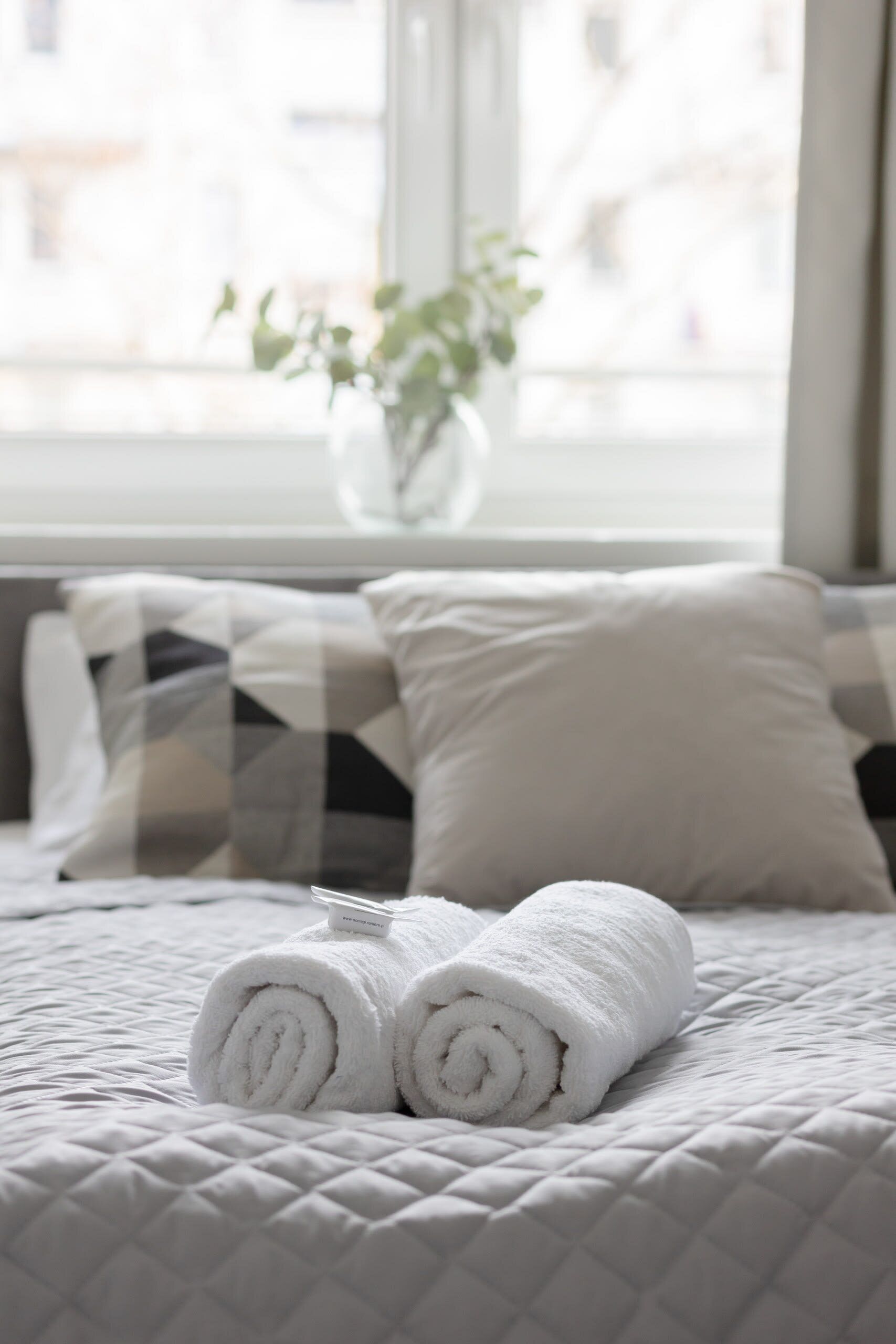 A neatly made bed with rolled white towels, a potted plant, and a window with a view of buildings.