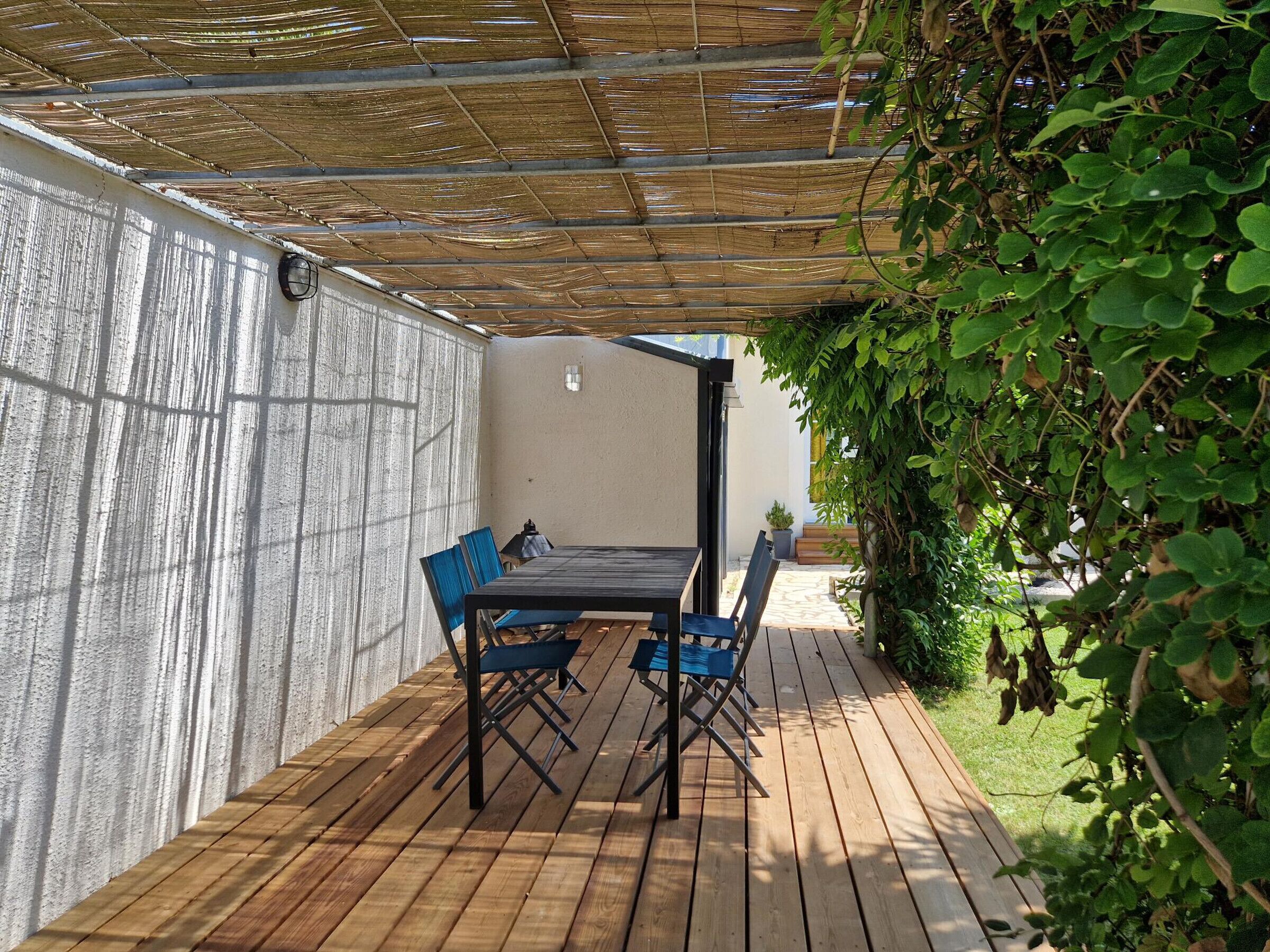 A covered patio with a wooden table and chairs, a white wall, and a garden area with green foliage.