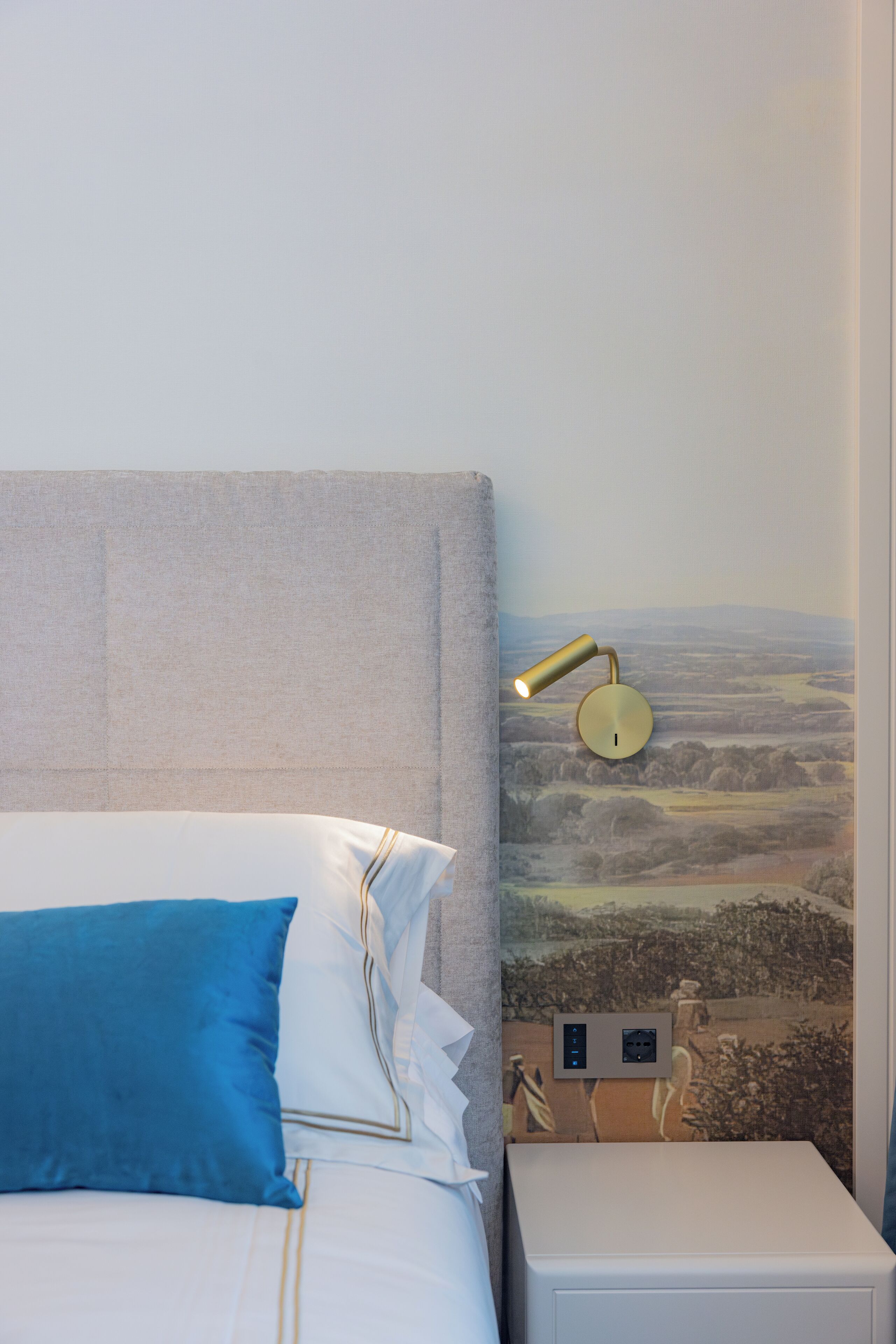 Ein ordentlich bezogenes Bett mit blauer und weißer Bettwäsche, eine Wandlampe sowie eine Steckdose an der Wand.