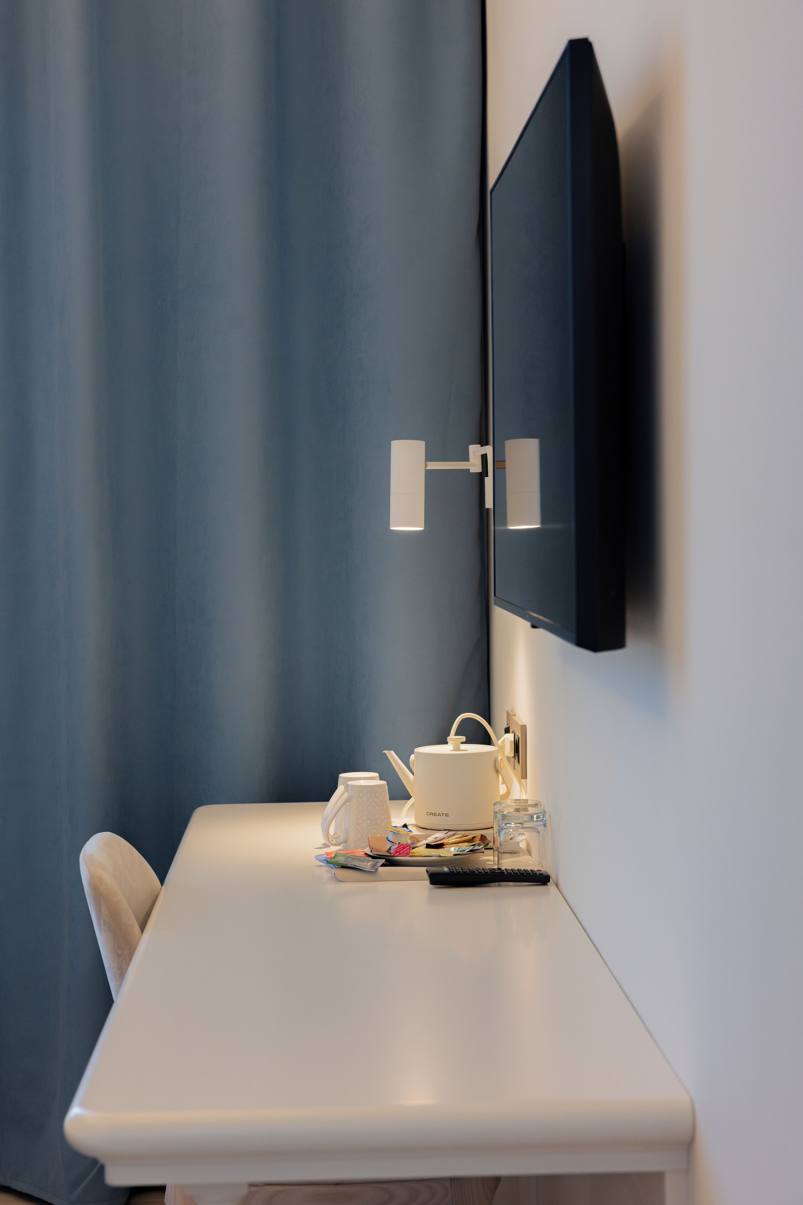 A modern hotel room with a white desk, a flat-screen TV mounted on the wall, a small table with a kettle and a cup, and a chair.