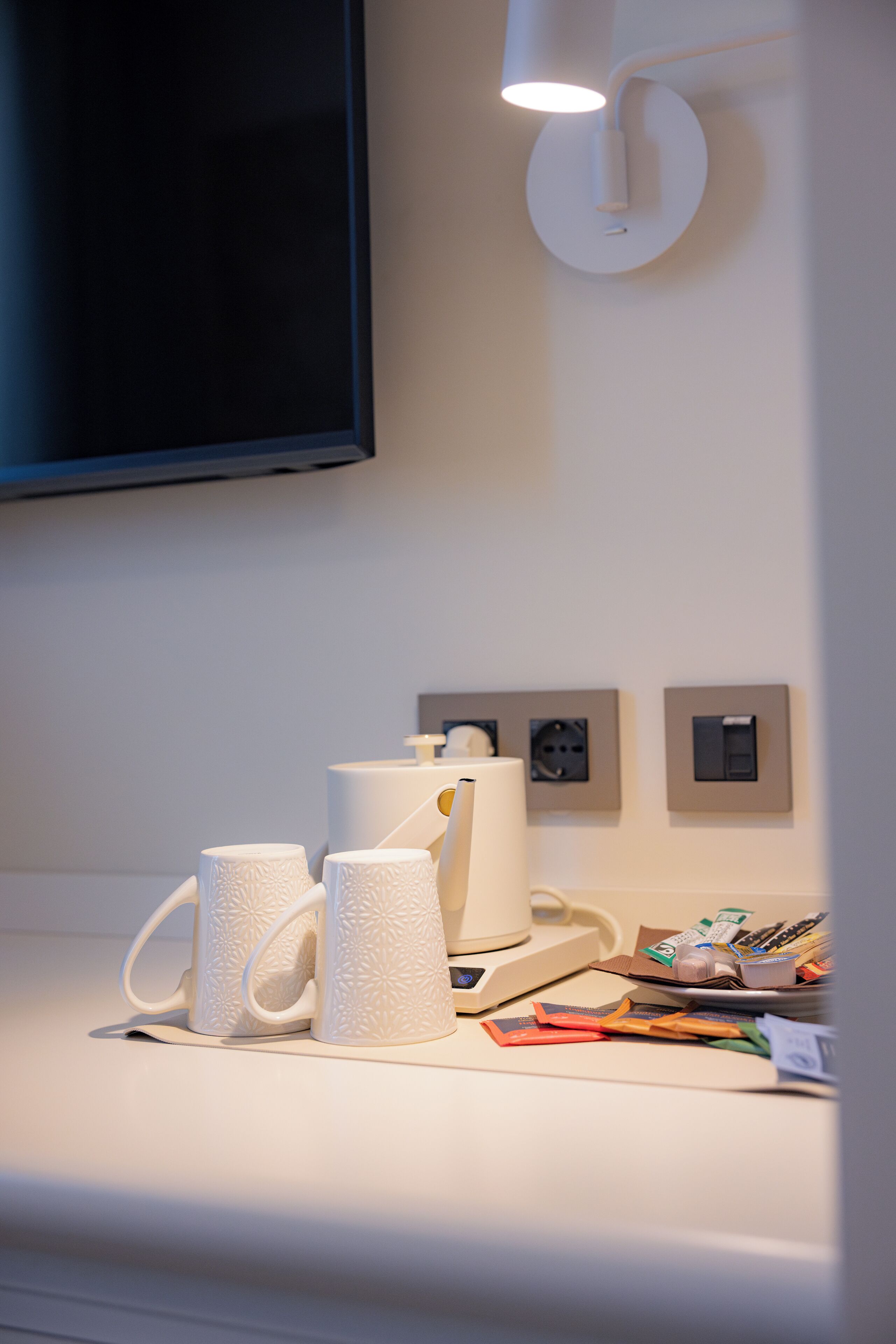 Ein sauberes, modernes Zimmer mit einem wandmontierten Flachbildfernseher, einem weißen Couchtisch mit zwei weißen Tassen, einer weißen Lampe und einigen verstreuten Gegenständen, darunter ein Telefon und ein paar Papiere.