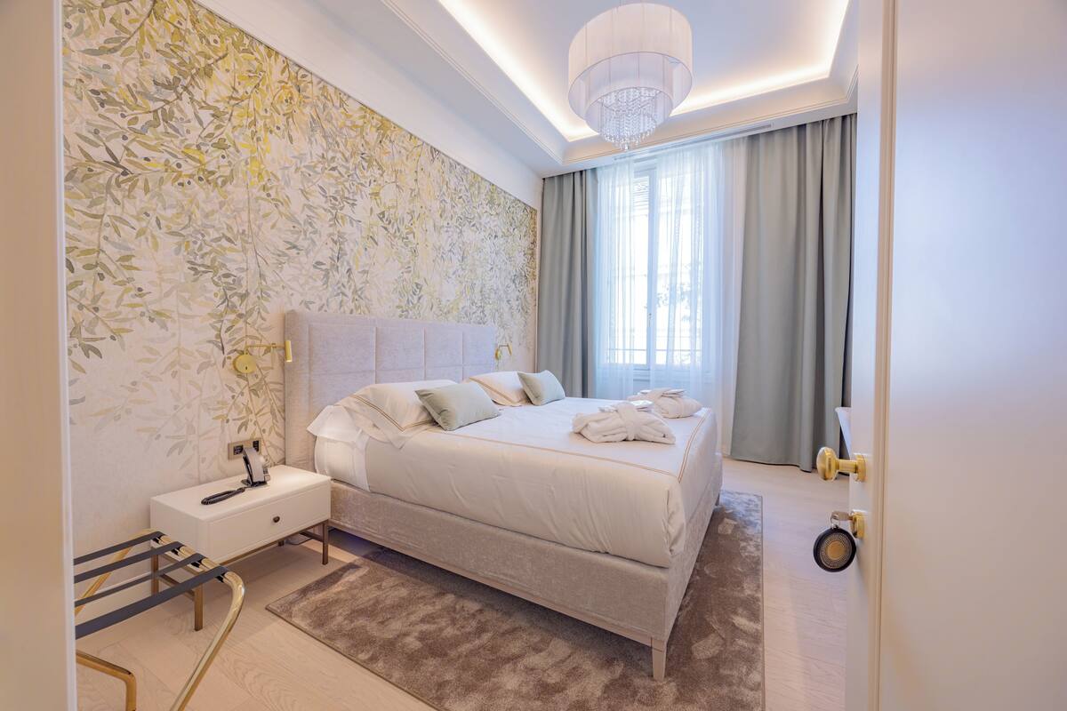 Une chambre à coucher moderne avec un grand lit, des tables de chevet, une commode, un lustre et une fenêtre avec des rideaux.