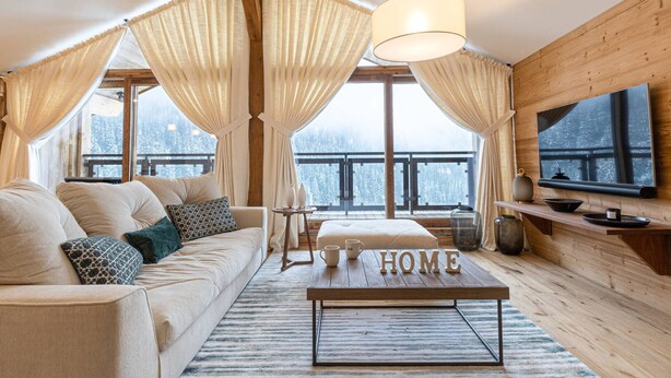 Une pièce chaleureuse avec un canapé, une table basse sur laquelle est inscrit le mot « MAISON  », un téléviseur fixé au mur et une vue sur des montagnes enneigées, visible à travers de grandes fenêtres.
