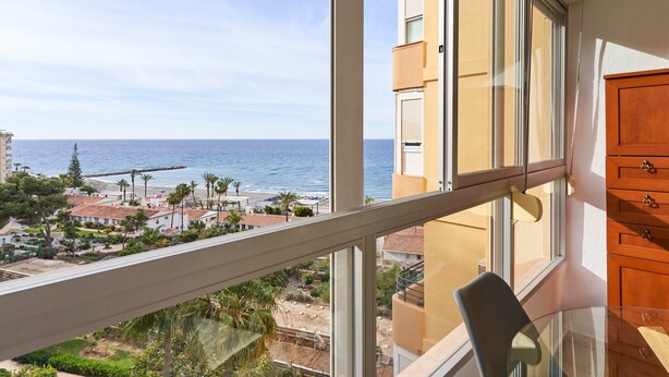 Un balcón con vista al mar, una mesa de vidrio y armarios de madera.