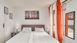 Una cama doble con cabecera, dos mesitas de noche con lámparas, un cuadro en la pared y una ventana con cortinas anaranjadas.