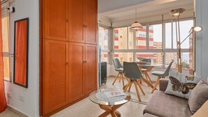 Una habitación con un armario de madera, una mesa de vidrio, un sofá con almohadas de mariposas y vistas a edificios a través de amplios ventanales.