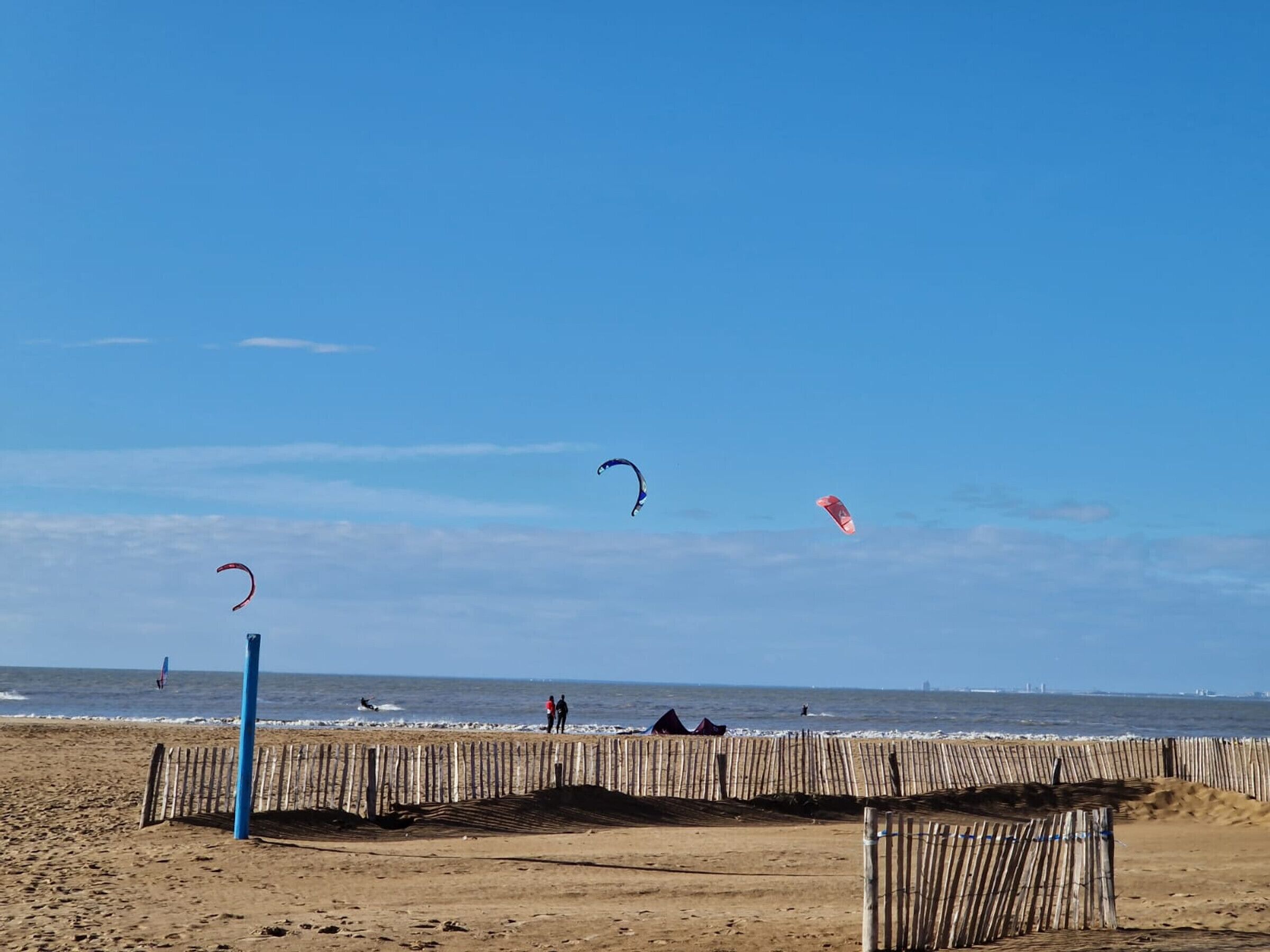 A beach with a wooden boardwalk, clear skies, and people flying kites.