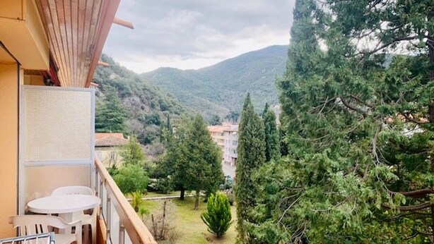 A balcony with a view of a mountainous landscape, a small garden, and a building in the distance.