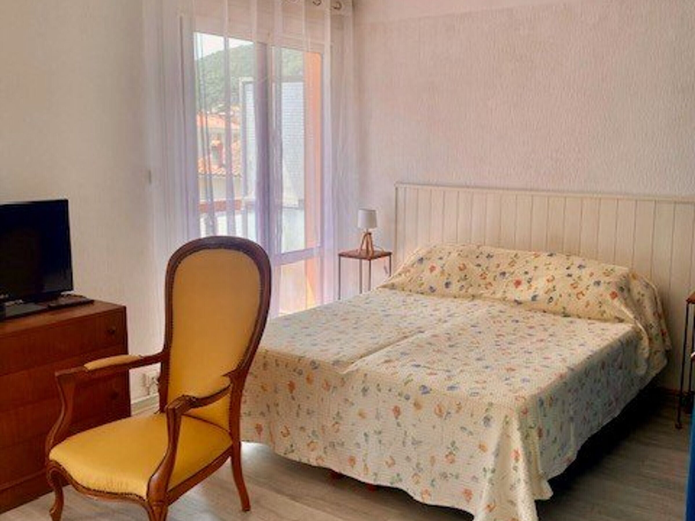 A bedroom with a bed, a chair, a TV on a wooden dresser, and a window with curtains.