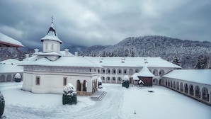 Un patio cubierto de nieve con un edificio central que tiene una cruz en la parte superior, rodeado de árboles nevados y un paisaje montañoso al fondo.