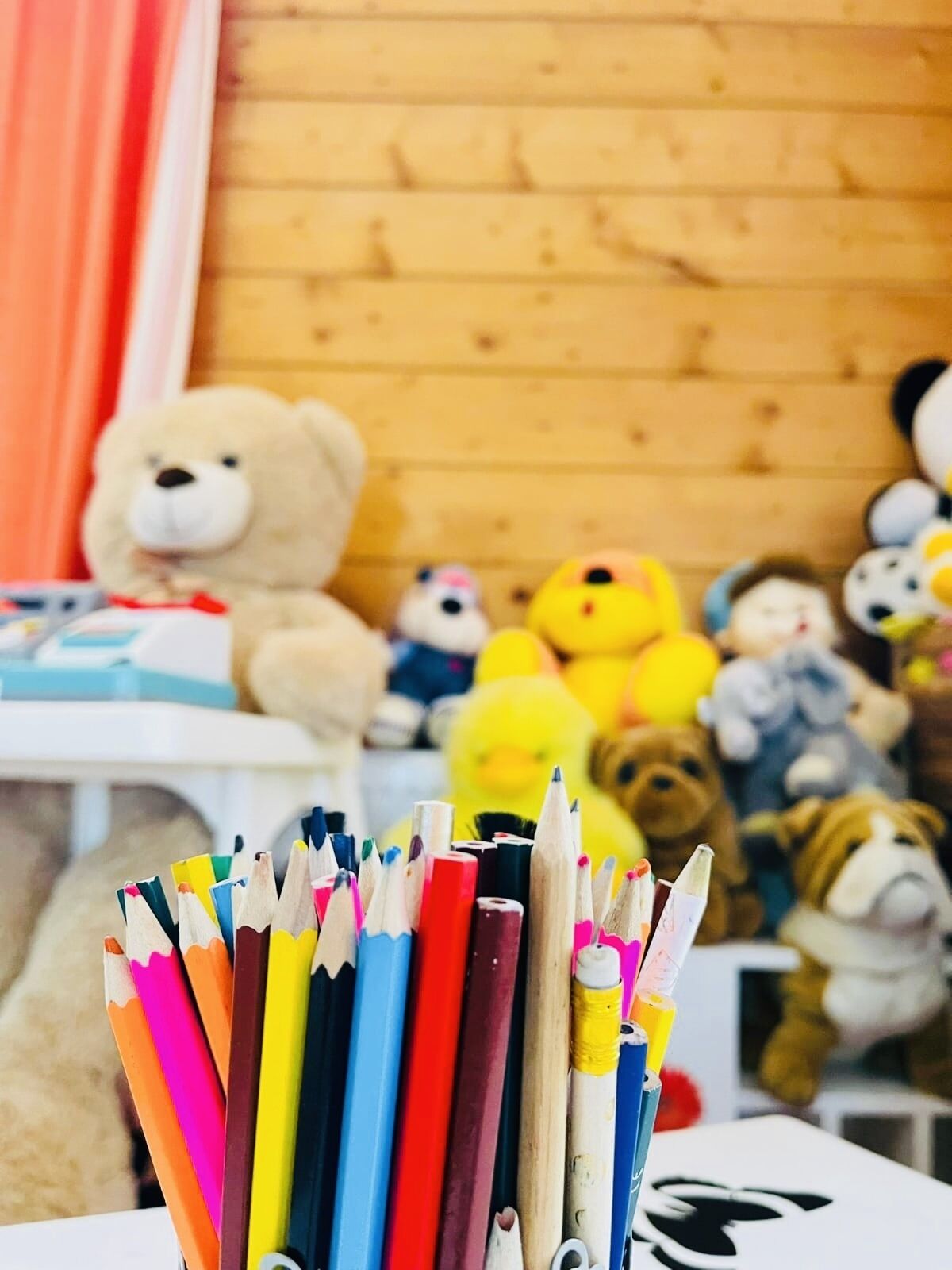 A collection of colored pencils in a holder, with various plush toys in the background.