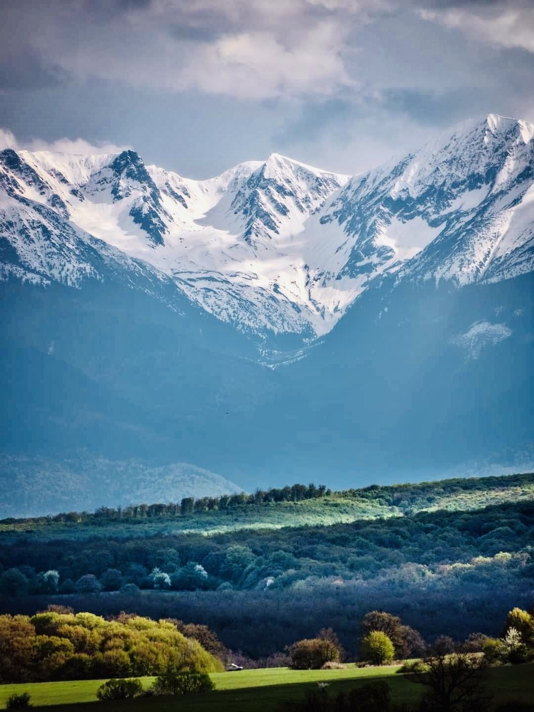 A mountainous landscape with snow-capped peaks and a valley with green fields and trees.