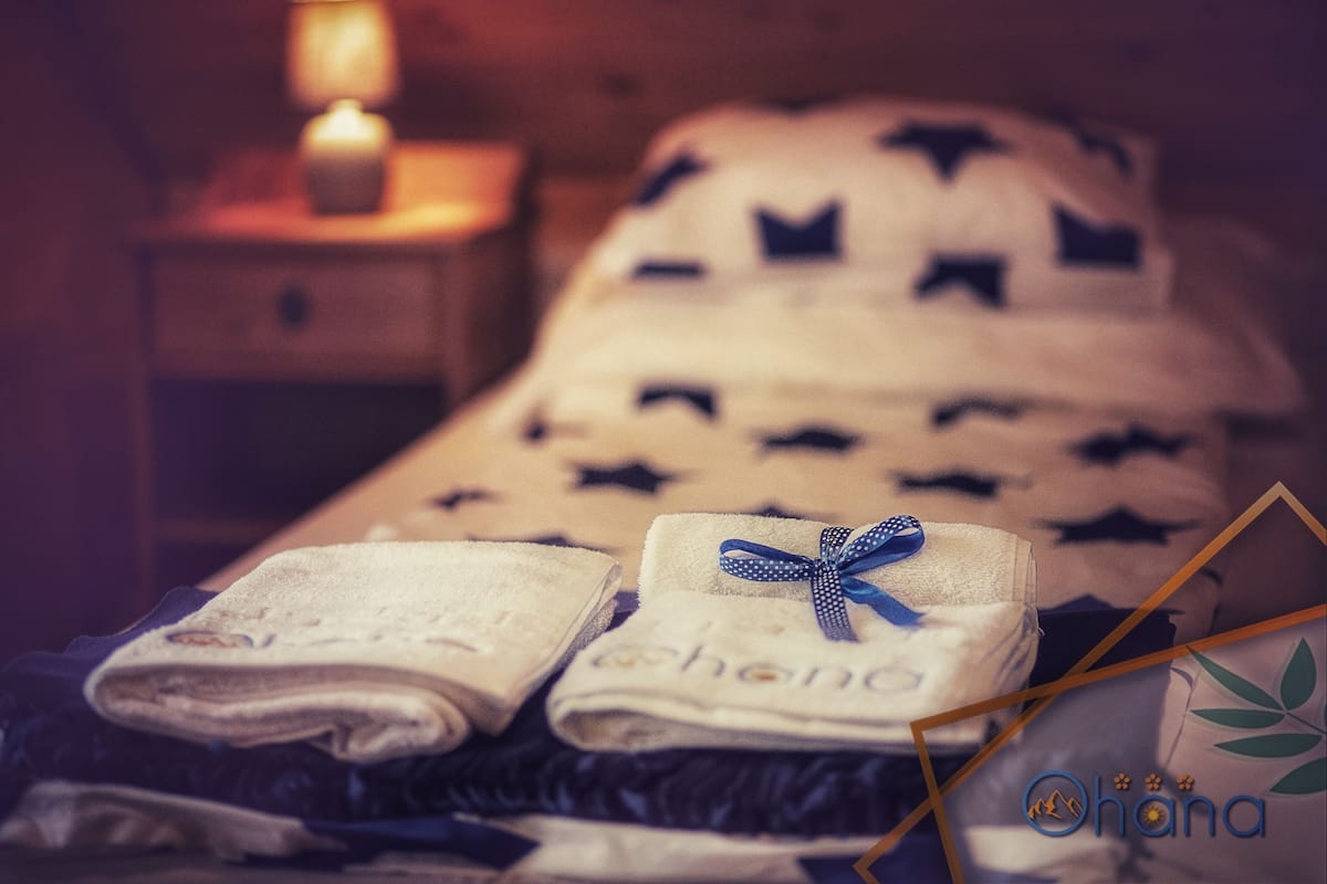 Una cama bien hecha con una manta blanca y un cojín estampado con motivos de estrellas. Sobre una mesita de noche, toallas dobladas adornadas con un lazo azul.
