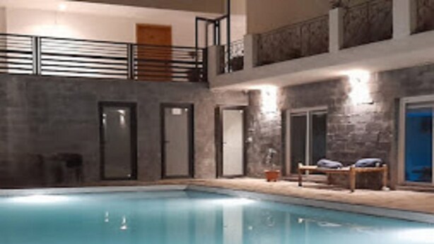 An indoor swimming pool with a stone-tiled floor, a wooden deck with lounge chairs, and a balcony with a railing. - Iris Lite Corbett (Ramnagar)