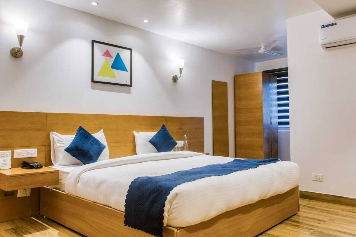 A modern hotel room with a wooden headboard, a large bed with white and blue bedding, a bedside table with a phone and charger, and a wall-mounted air conditioner.