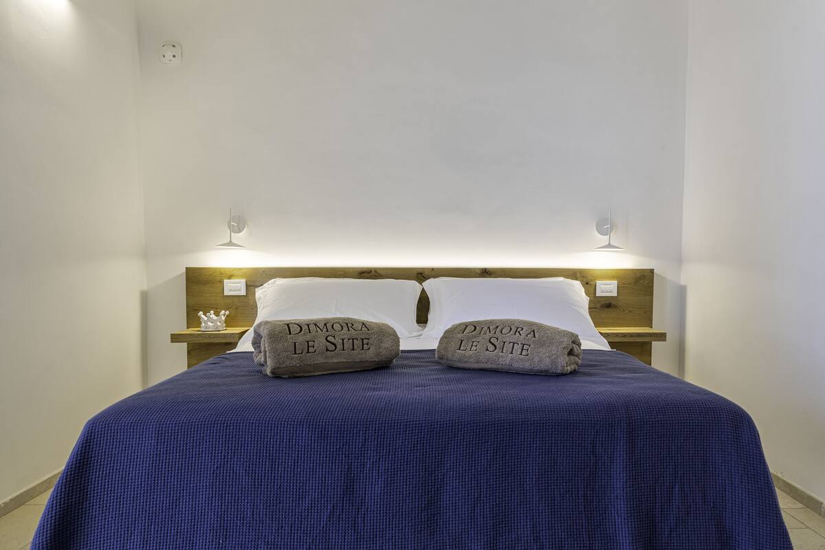 A neatly made bed with a blue bedspread, two pillows with "Dimora Le Site" text, and bedside lamps.