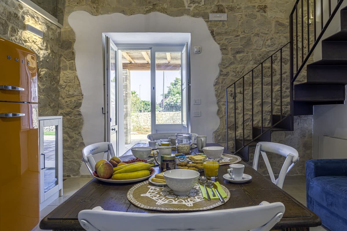 A dining area with a stone wall, a wooden table set with breakfast items, and a view of the outdoors.