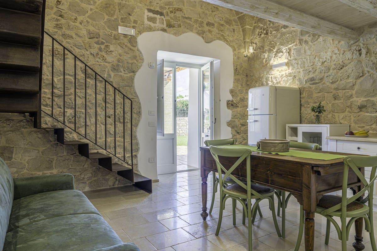 A cozy interior with a stone wall, a wooden staircase, a dining table with chairs, and a kitchen area.